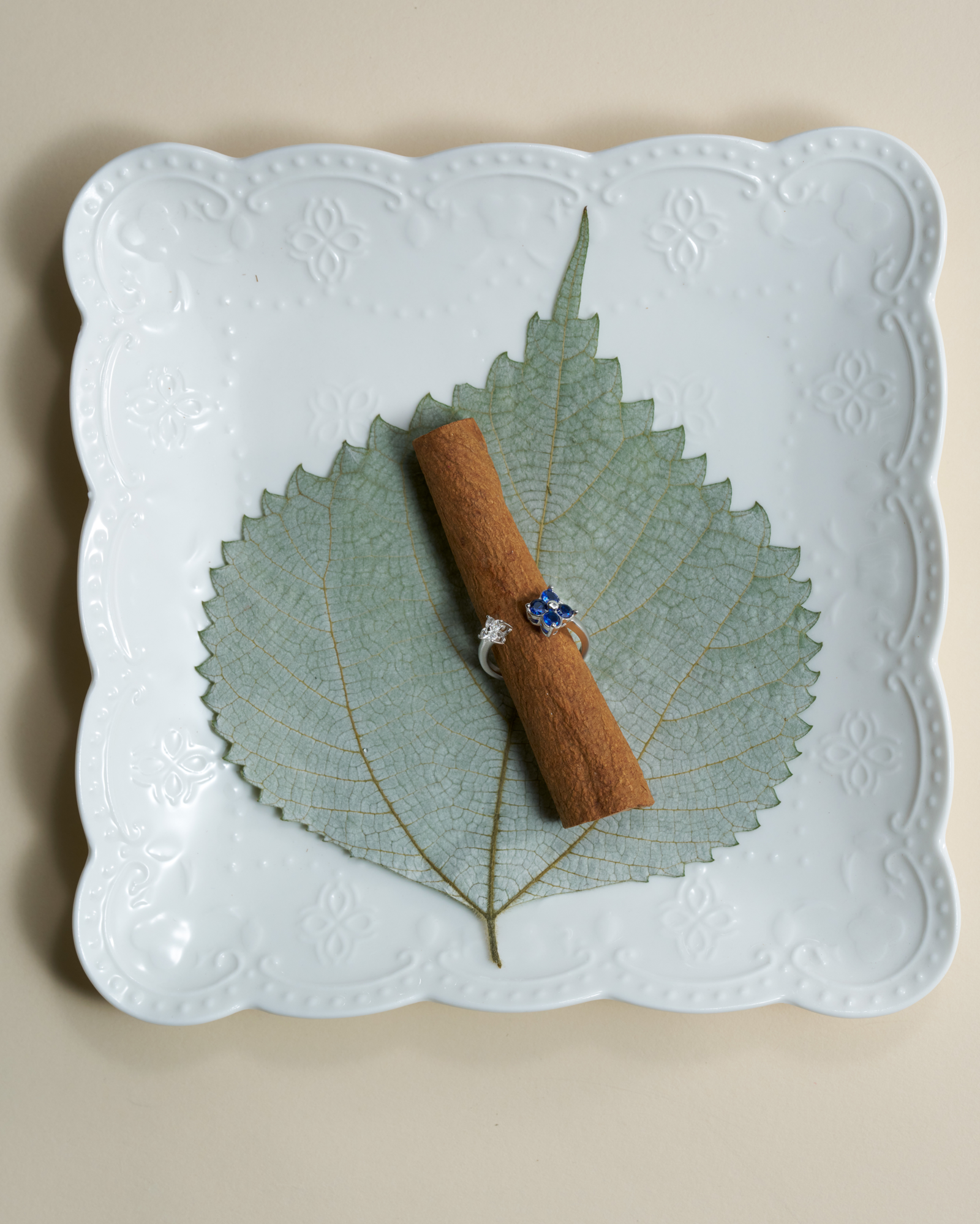 A silver ring with blue and white patterned flowers is displayed on a small cinnamon stick, which rests on a green leaf atop a white ceramic plate against a beige background.