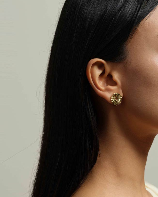 A close-up side profile of a woman with long black hair wearing a textured gold flower-shaped stud earring against a neutral light gray background.