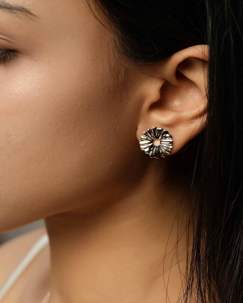 A close-up of a model's ear wearing a single silver-toned flower-shaped stud earring with a textured, petal-like design.
