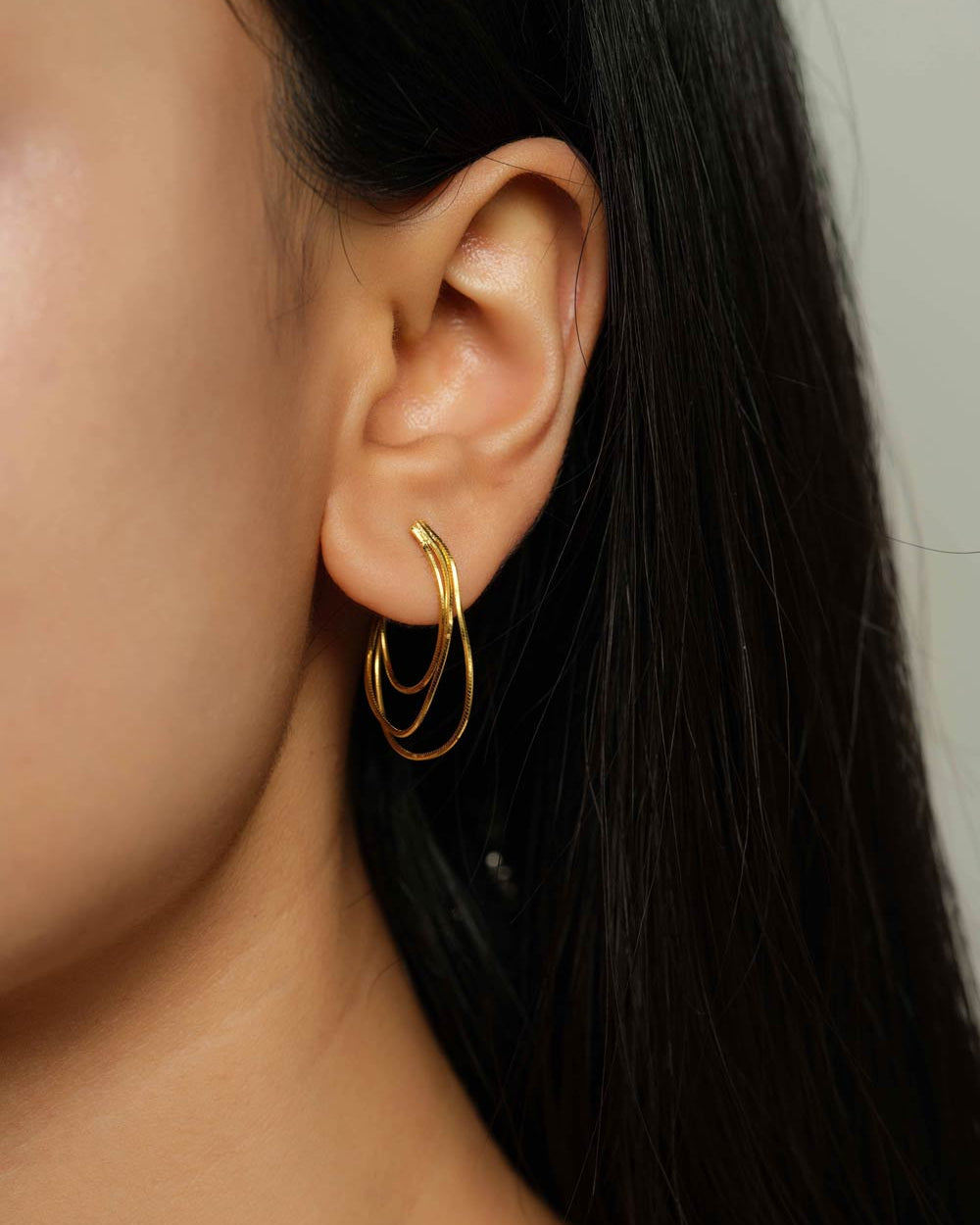 A close-up of a model's ear wearing a pair of gold hoop earrings, with her dark hair and skin visible against a soft neutral background.