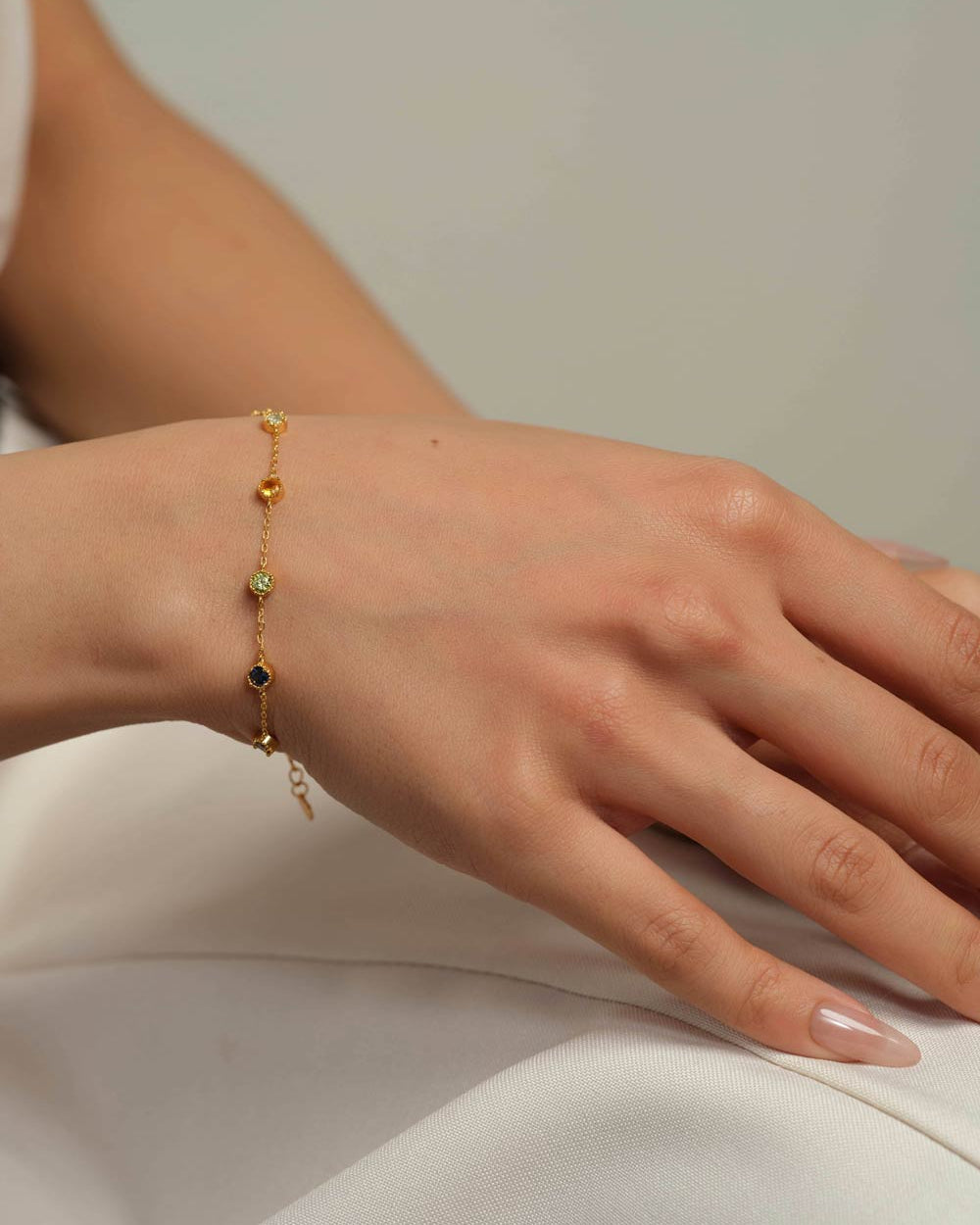 A delicate gold chain bracelet with small colorful gemstone accents, shown worn on a person's wrist against a soft neutral background.