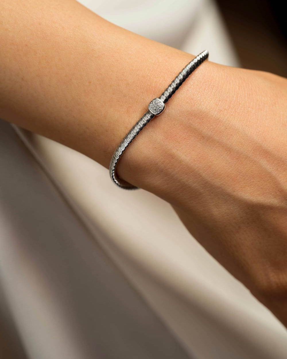 A close-up of a person's wrist wearing a slim silver-toned bracelet with a textured band and a central round pavé-set stone.