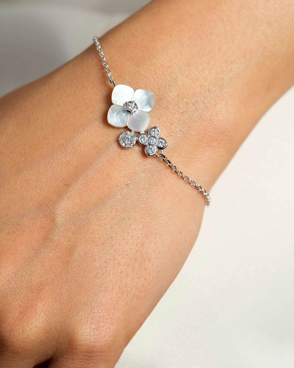A delicate silver chain bracelet worn on a wrist, featuring two flower charms: one made of white mother-of-pearl petals with a clear crystal center, and another small flower cluster set with clear crystals.