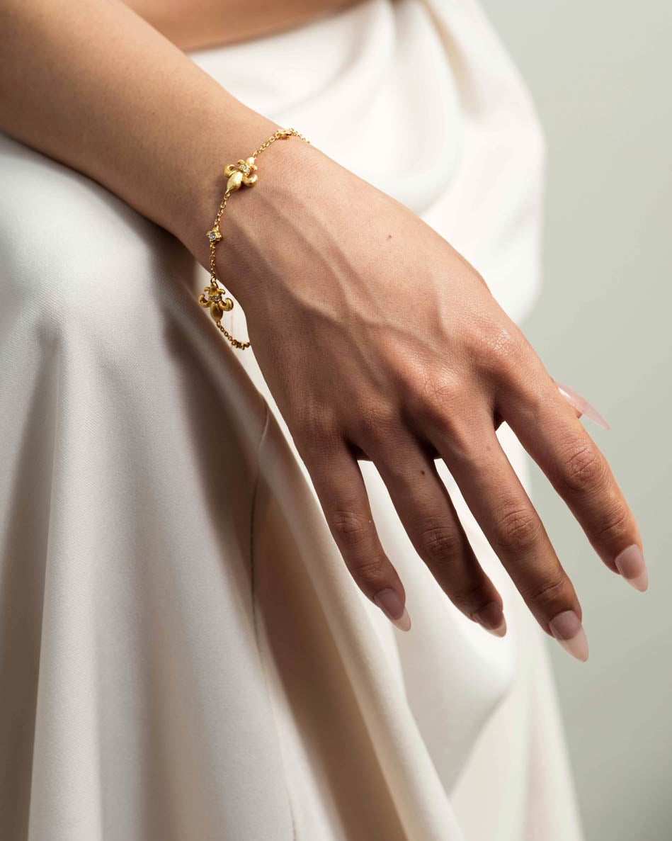 A delicate gold chain bracelet with small floral or irsh-like charms worn on a woman's wrist, shown against a soft cream dress and pale background.