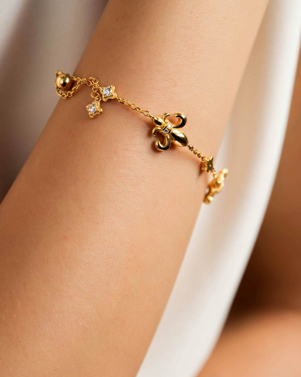 A delicate gold chain bracelet with three dangling charms, including a fleur-de-lis and two small gold beads, shown worn on a person's wrist.