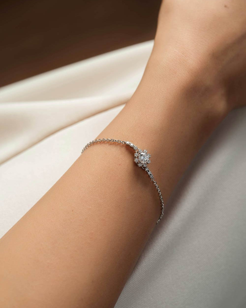 A delicate silver-toned bracelet with a central cluster of clear stones, shown worn on a person's wrist against a soft cream fabric background.