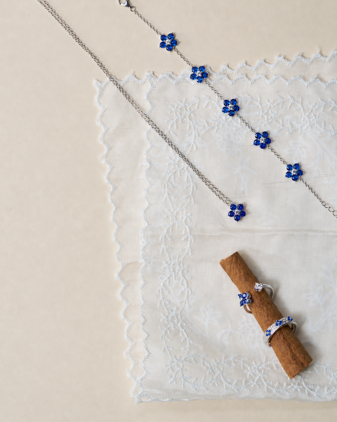 Silver chain jewelry set with blue flower charms, displayed on a white lace cloth with a cinnamon stick accent on a beige background.
