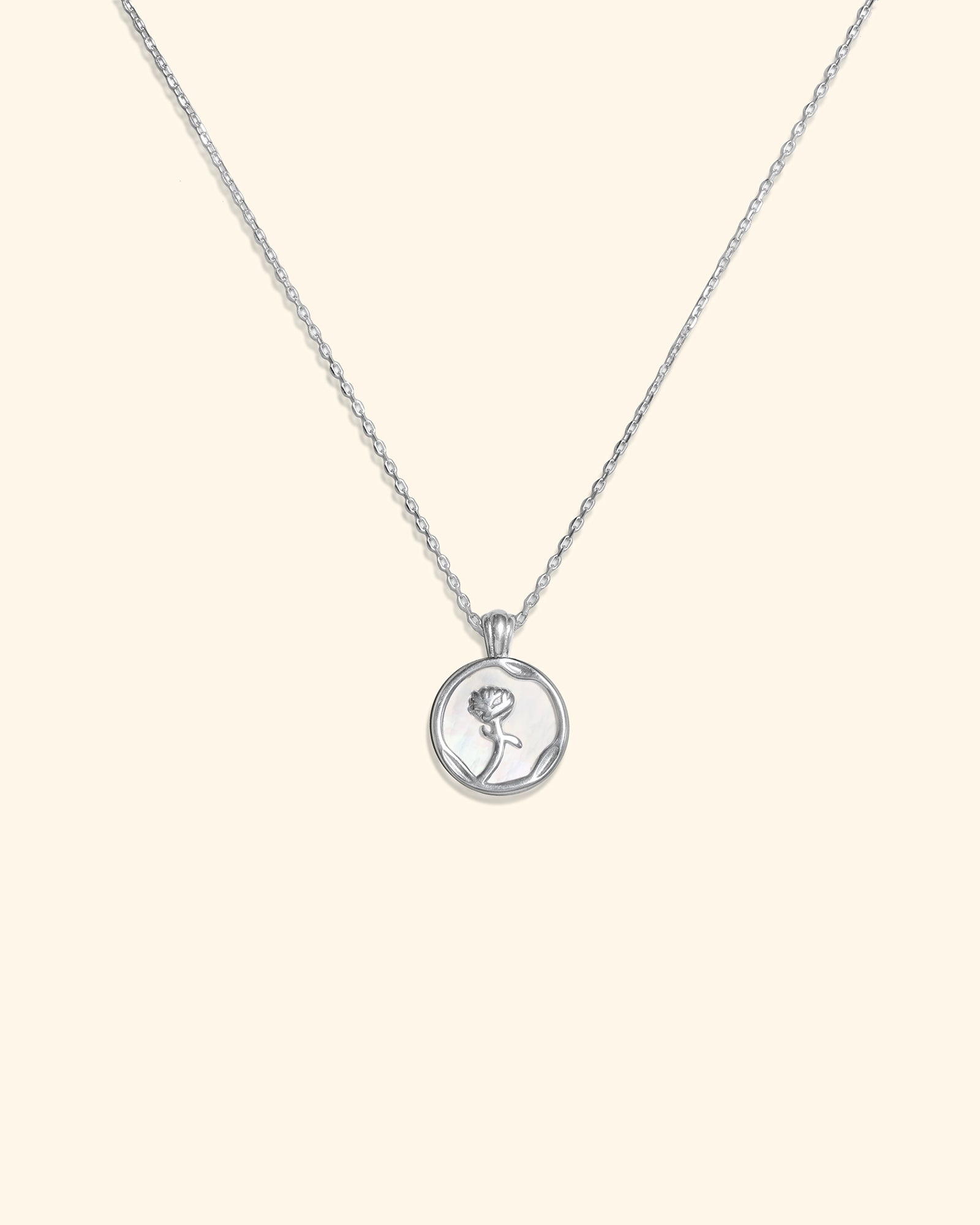 A silver chain necklace with a small round pendant featuring an engraved silhouette of a rose, displayed against a solid cream background.