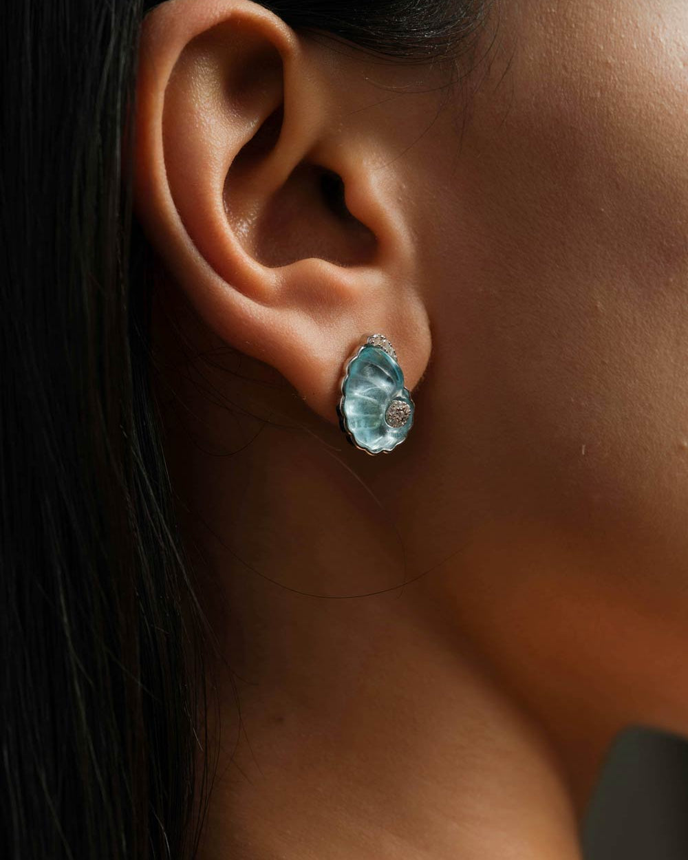 A close-up side profile of a woman's ear wearing a shell-shaped light blue crystal stud earring with silver setting.