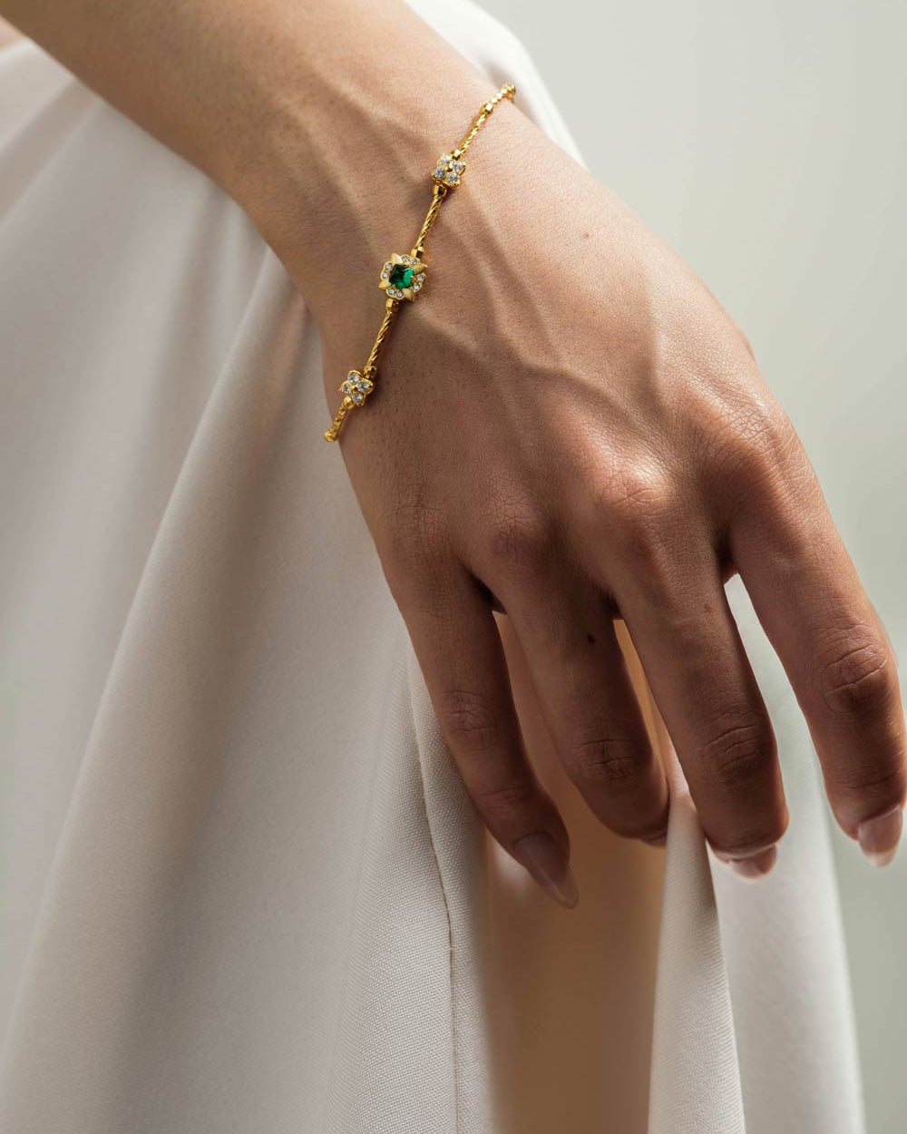 A delicate gold chain bracelet with green and clear gemstones worn on a woman's wrist, shown against a soft white dress and a light gray background.