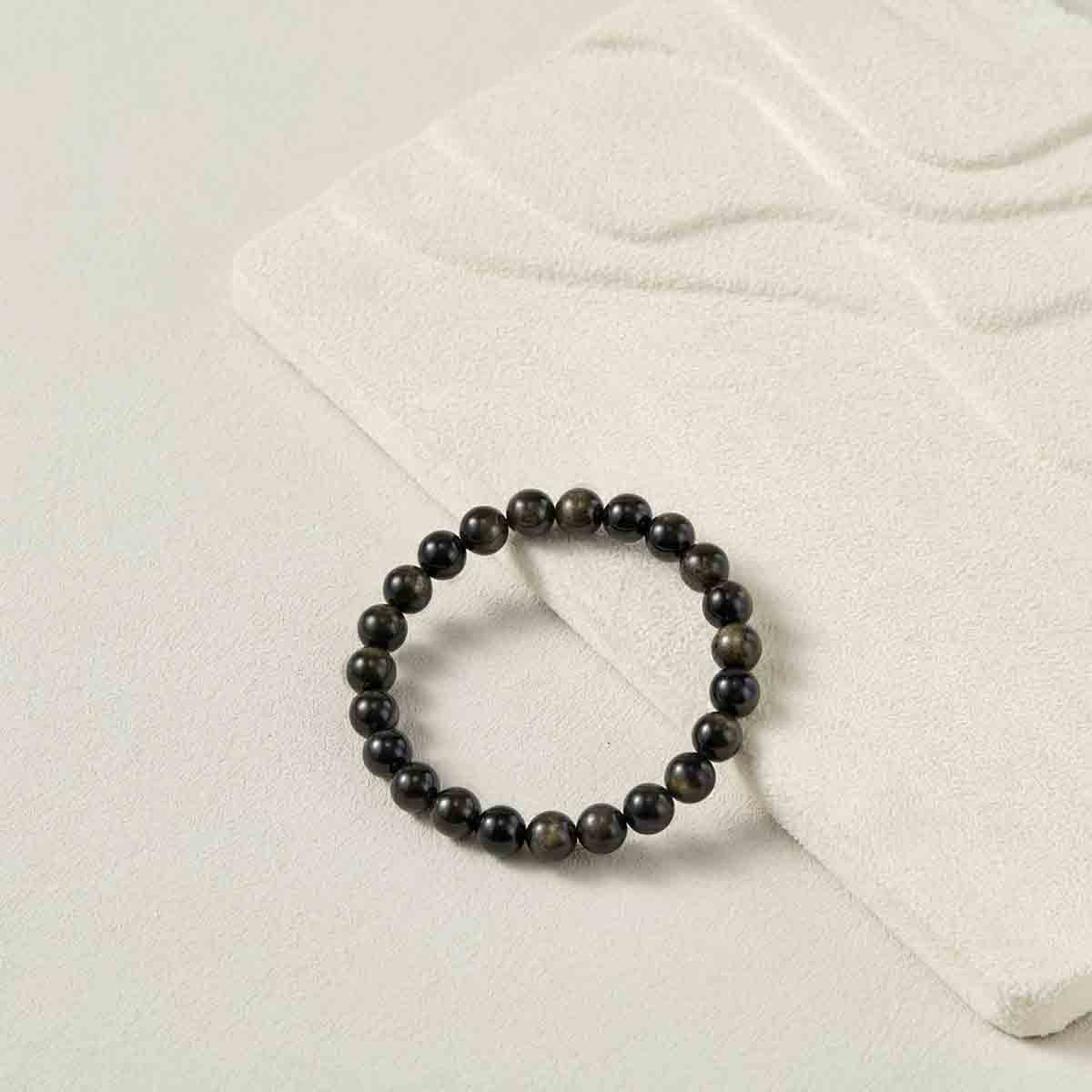 Still life photography of Choodo pure natural Gold Sheen Obsidian crystal beaded bracelet laid flat on an oatmeal velvet jewelry display board, capturing the minimalist premium textures.