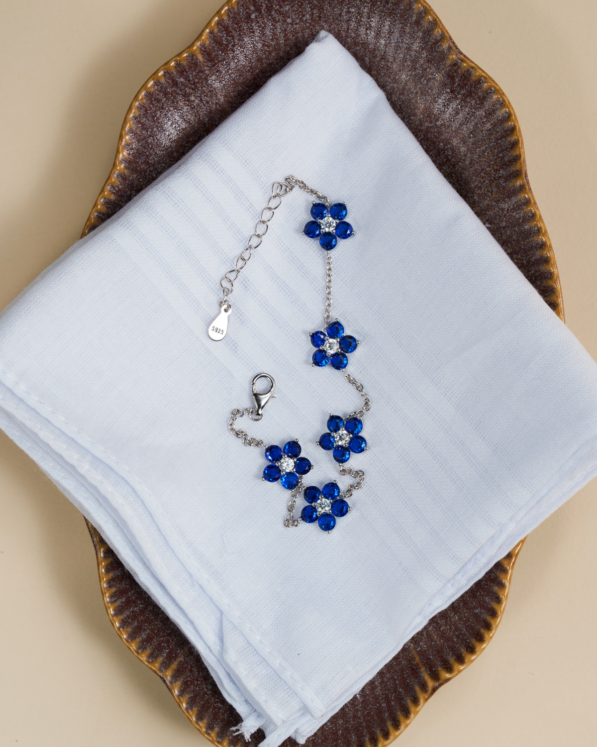A silver chain bracelet with blue flower charms, displayed on a white folded cloth over a brown scalloped plate against a beige background.