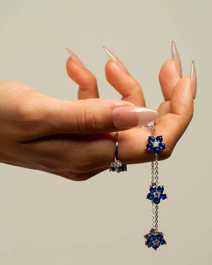 A close-up of a hand with long nude manicured nails holding a pair of dangling earrings. The earrings feature silver-toned chains and floral clusters set with deep blue stones, with a small silver ring also visible on the hand.