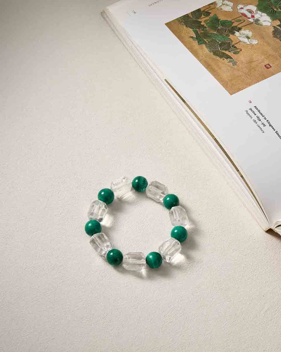 Still life photography of Choodo natural Malachite and Clear Quartz geometric bracelet laid flat on a beige tabletop next to a book, minimalist intellectual aesthetic