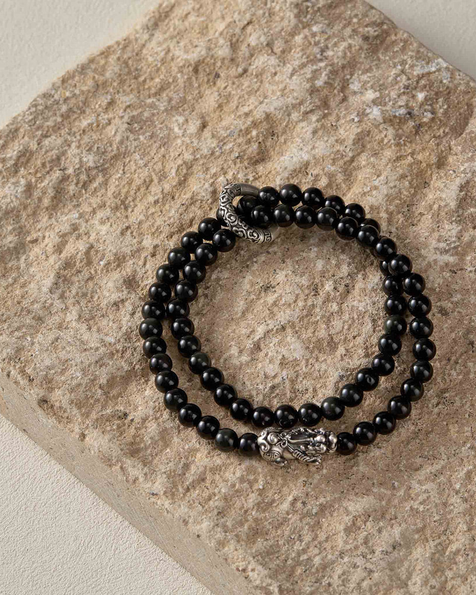 Still life photography of Choodo men's natural Obsidian bracelet with silver Pixiu laid flat on a rustic natural stone board, organic masculine textures contrast.