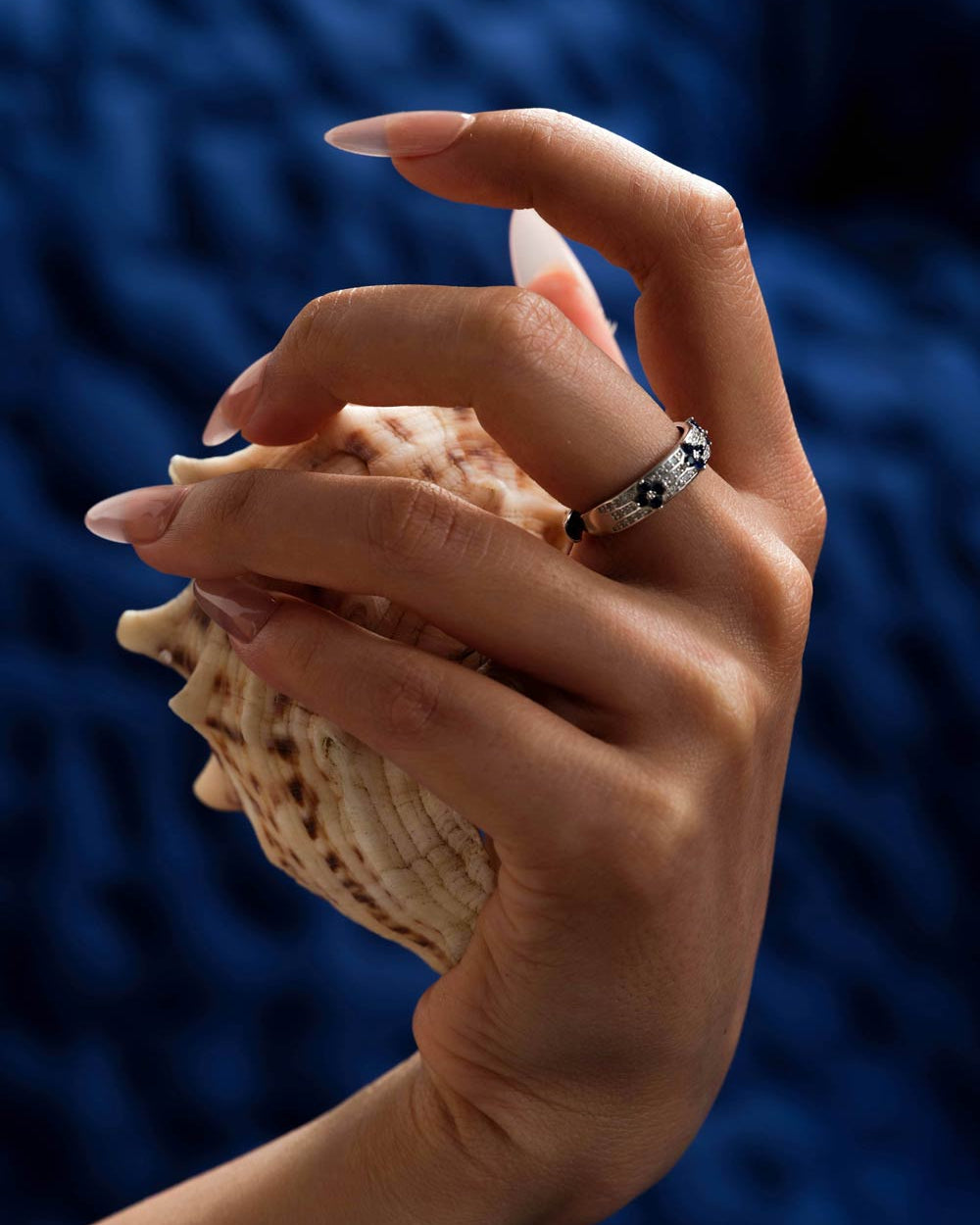A close-up of a hand holding a seashell, wearing a silver band ring with black and white floral details, set against a dark blue textured background.