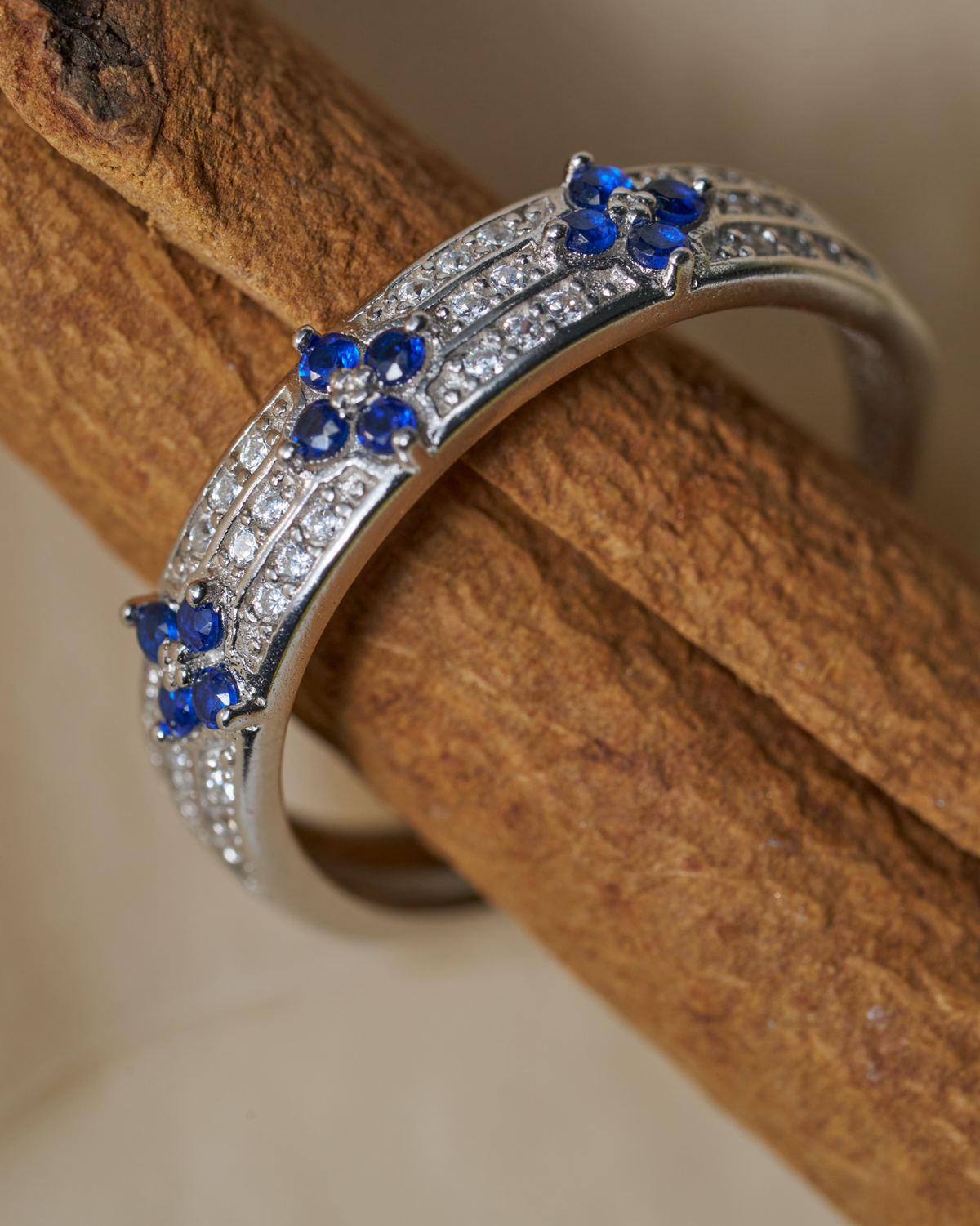 A silver-toned curved ring with rows of small clear stones and three blue gemstone floral motifs, displayed on a textured brown wooden stick against a soft beige background.
