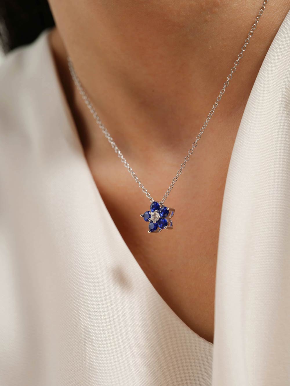 A close-up of a woman wearing a delicate silver chain necklace with a small flower-shaped pendant featuring deep blue petals and a clear center stone, styled with a white satin top.