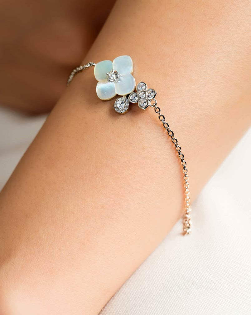 A delicate chain bracelet with mother of pearl flower charms worn on a wrist. One flower charm is made of milky white mother-of-pearl, while the other is set with small clear crystals.