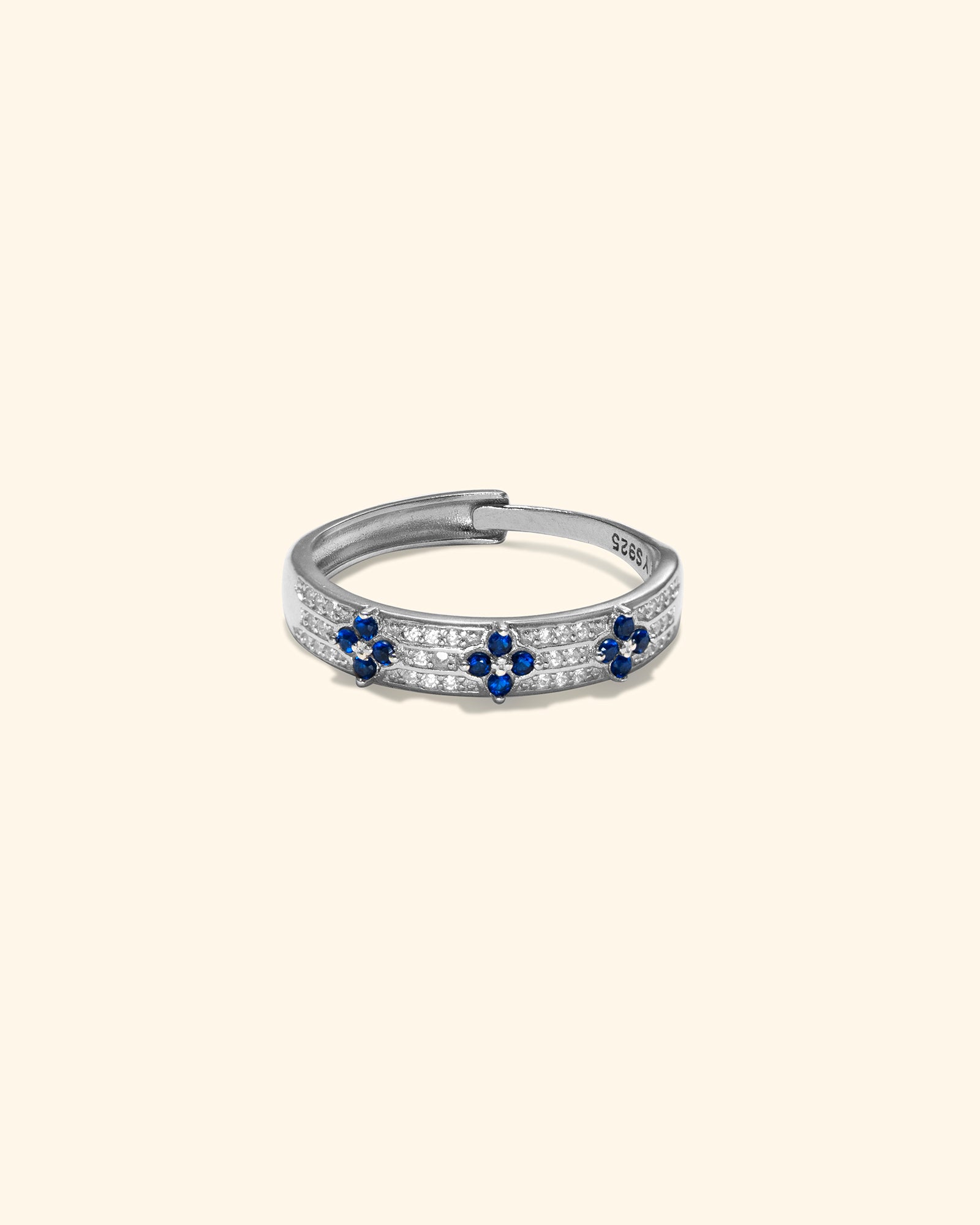 A silver-toned hinged ring featuring rows of small clear stones and three blue flower motifs, set against a solid pale cream background.