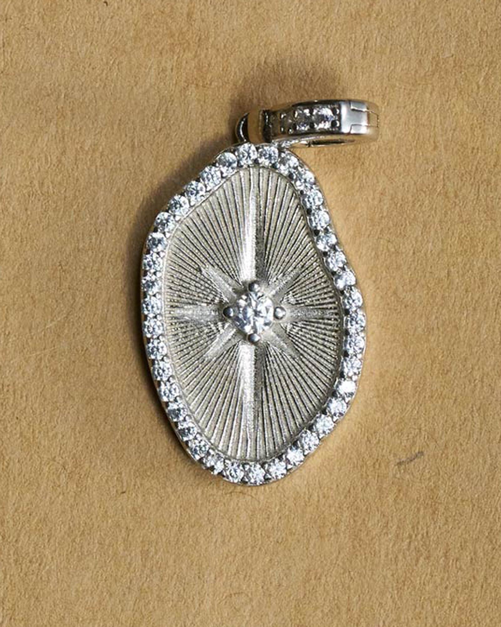 A silver-toned oval-shaped pendant with a starburst center and a border of small clear stones, displayed on a textured tan background.
