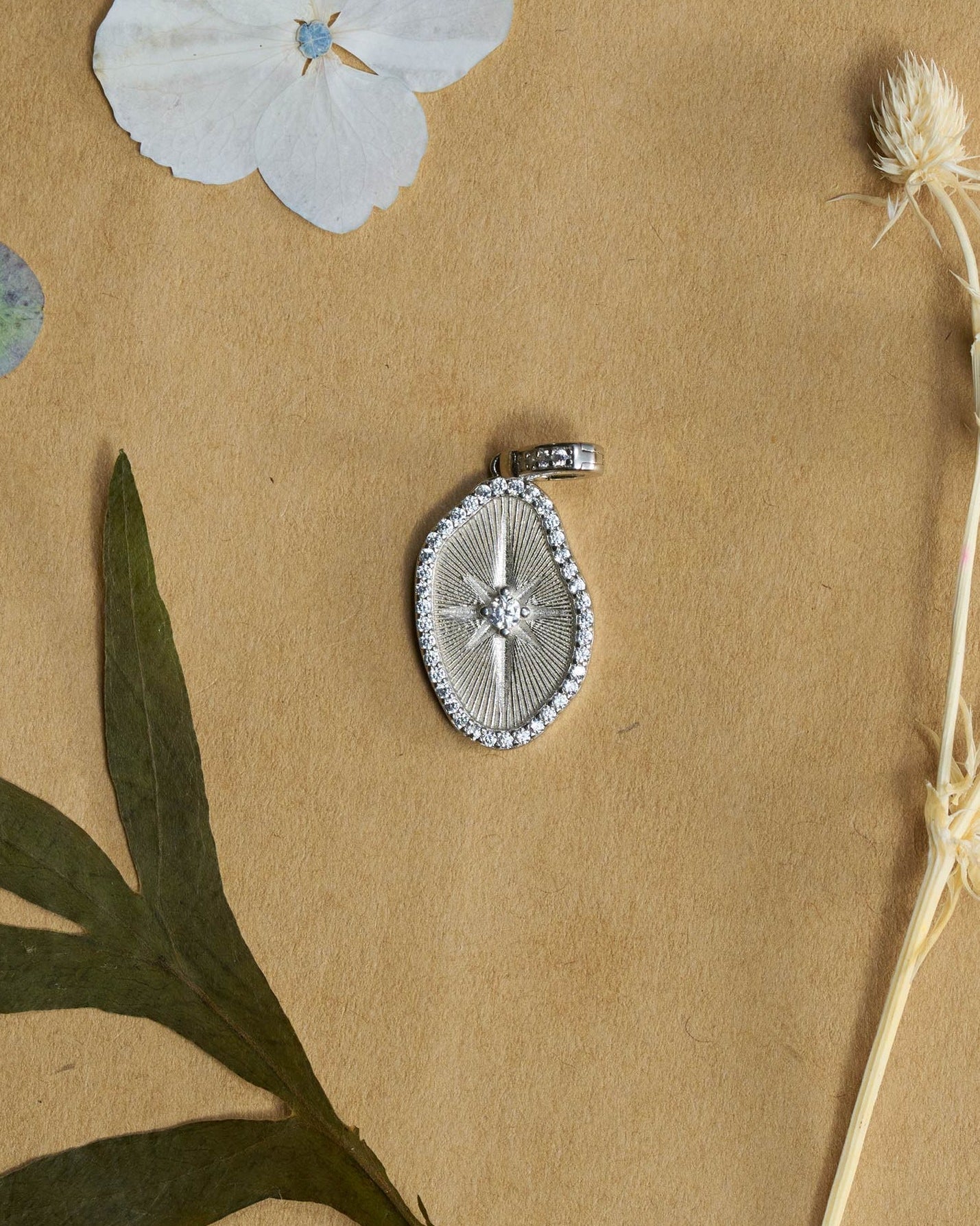 A silver oval-shaped pendant with a starburst center and a beaded border, displayed on a textured brown paper background with dried flowers and green leaves.