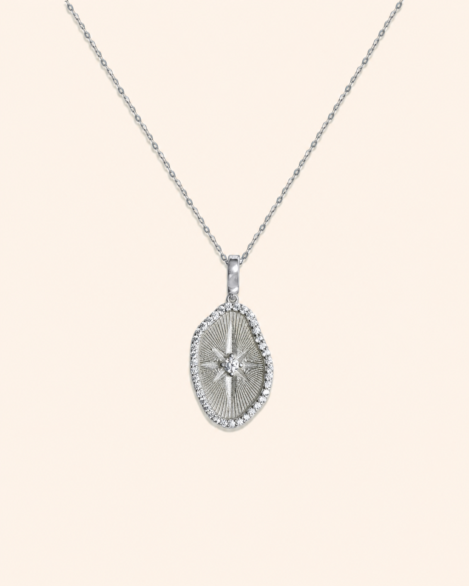 A silver pendant necklace featuring a textured oval pendant with a central starburst motif and a border of small zircons, suspended from a delicate silver chain against a light beige background.
