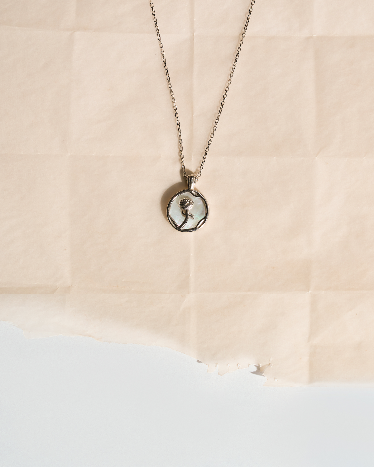 A silver-toned chain necklace with a round pendant featuring a black-and-white portrait of a woman, displayed on a torn sheet of beige paper against a white background.