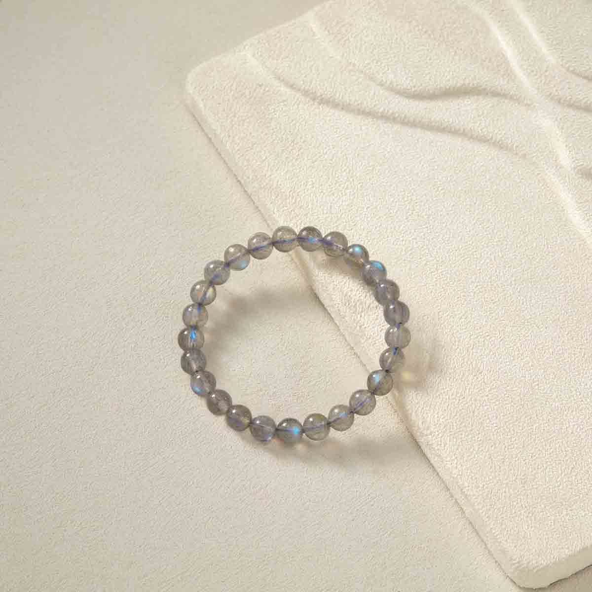 Still life photography of Choodo pure natural Veil Grey Moonstone crystal beaded bracelet laid flat on an oatmeal velvet jewelry display board, capturing the minimalist premium textures.