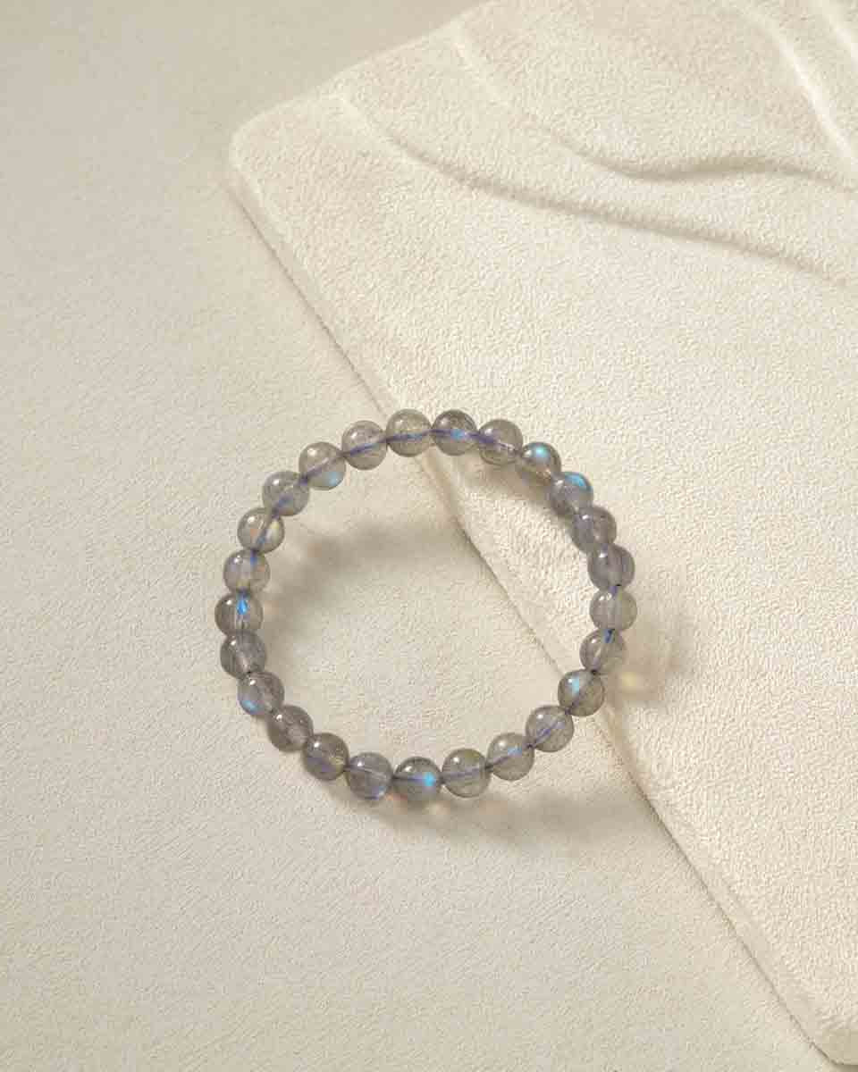 Still life photography of Choodo pure natural Veil Grey Moonstone crystal beaded bracelet laid flat on an oatmeal velvet jewelry display board, capturing the minimalist premium textures.