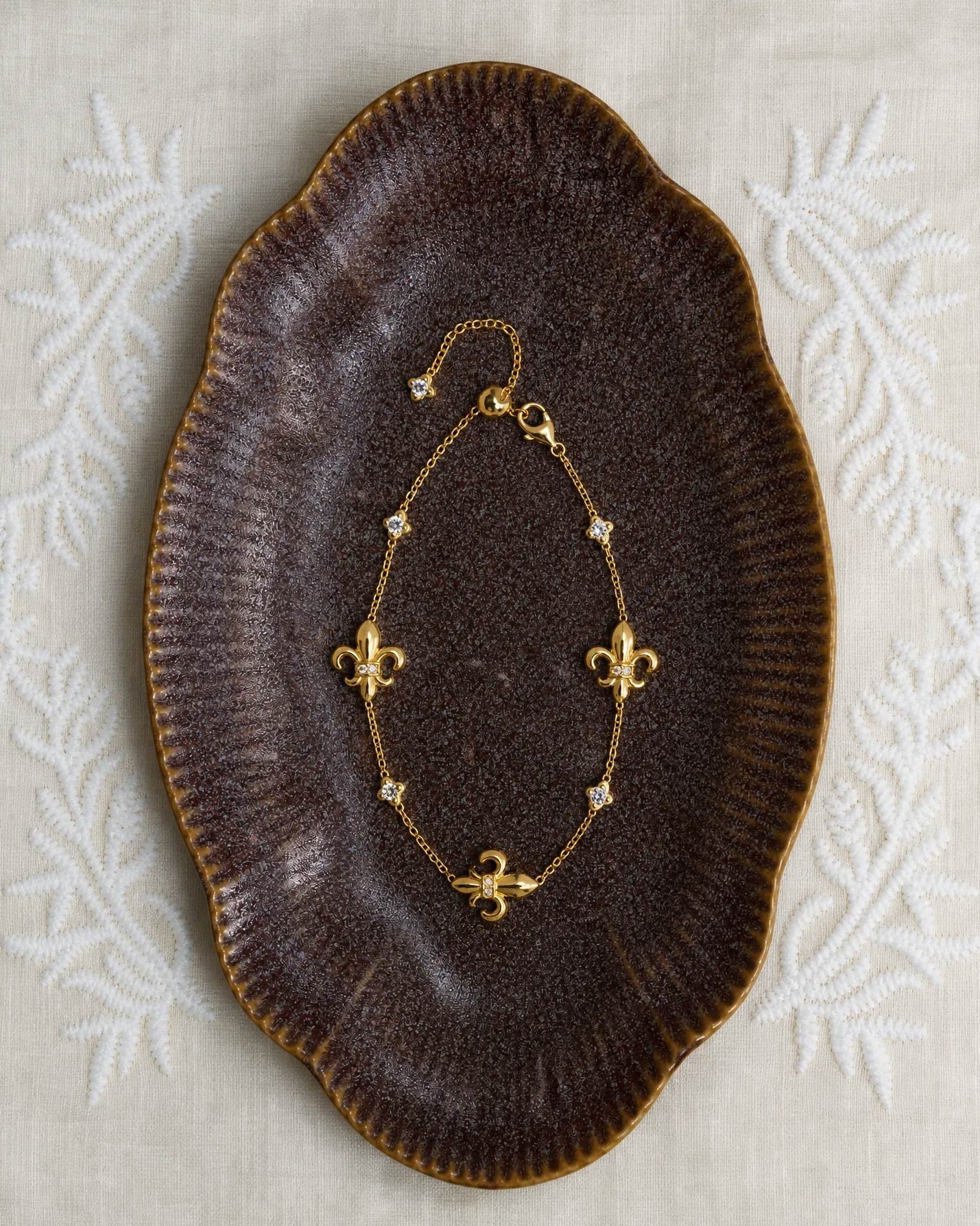 A delicate gold chain bracelet with fleur-de-lis charms, displayed on a dark brown ceramic dish over a light beige embroidered fabric background.