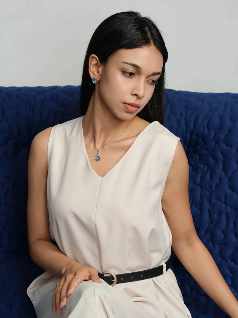 A woman with long black hair sits on a textured blue couch wearing a sleeveless cream-colored top and white pants with a black belt. She is wearing a small blue teardrop pendant necklace and matching blue earrings.
