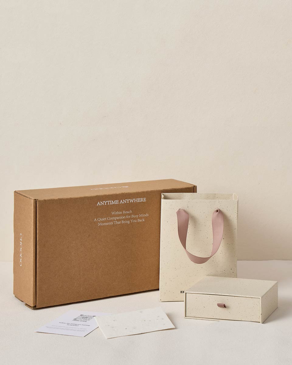 A kraft brown gift box with white text, a cream paper shopping bag with dusty pink handles, a small cream drawer-style gift box, and a few cards on a light beige background.