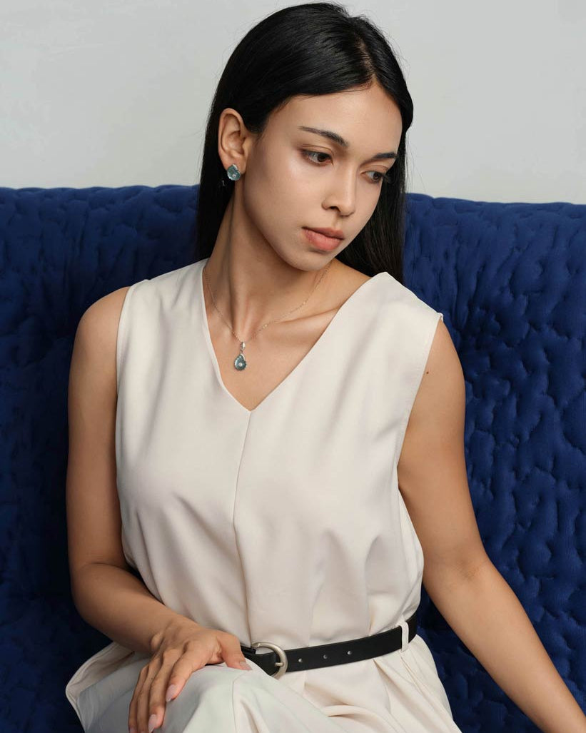 A woman with long black hair sits on a textured blue couch wearing a sleeveless cream-colored top and white pants with a black belt. She is wearing a small blue shell pendant necklace and matching blue earrings.