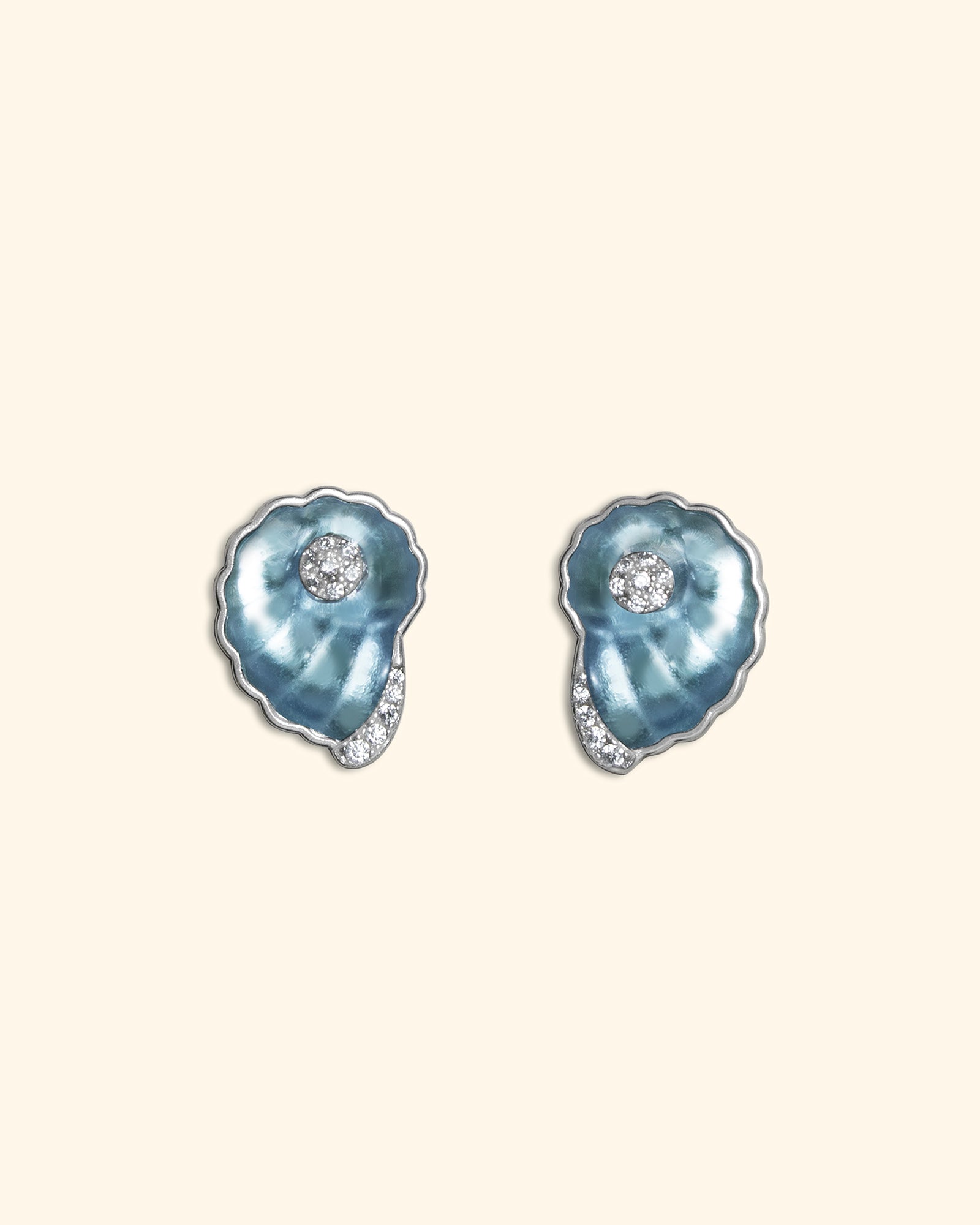 A pair of ocean blue shell stud ear studs with small clear crystal accents, shown on a solid cream background.