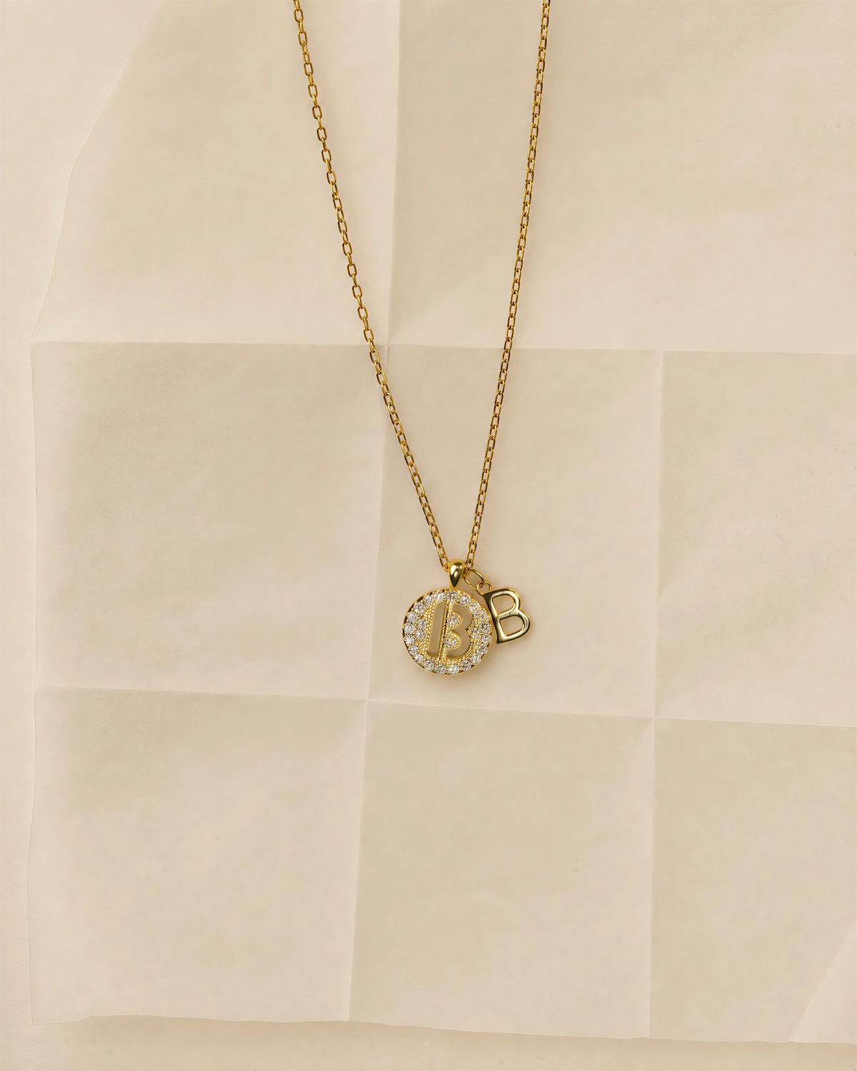 Choodo 18k gold plated sterling silver dual 'B' letter pendant necklace placed on translucent beige frosted paper