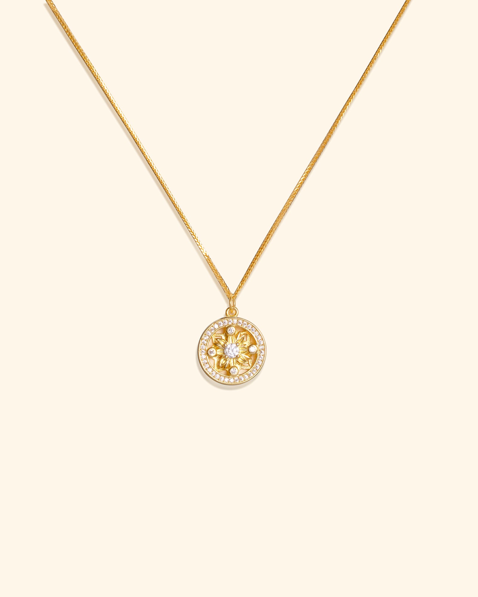 A gold chain necklace with a circular pendant featuring a gold flower motif surrounded by small clear stones, set against a solid cream background.