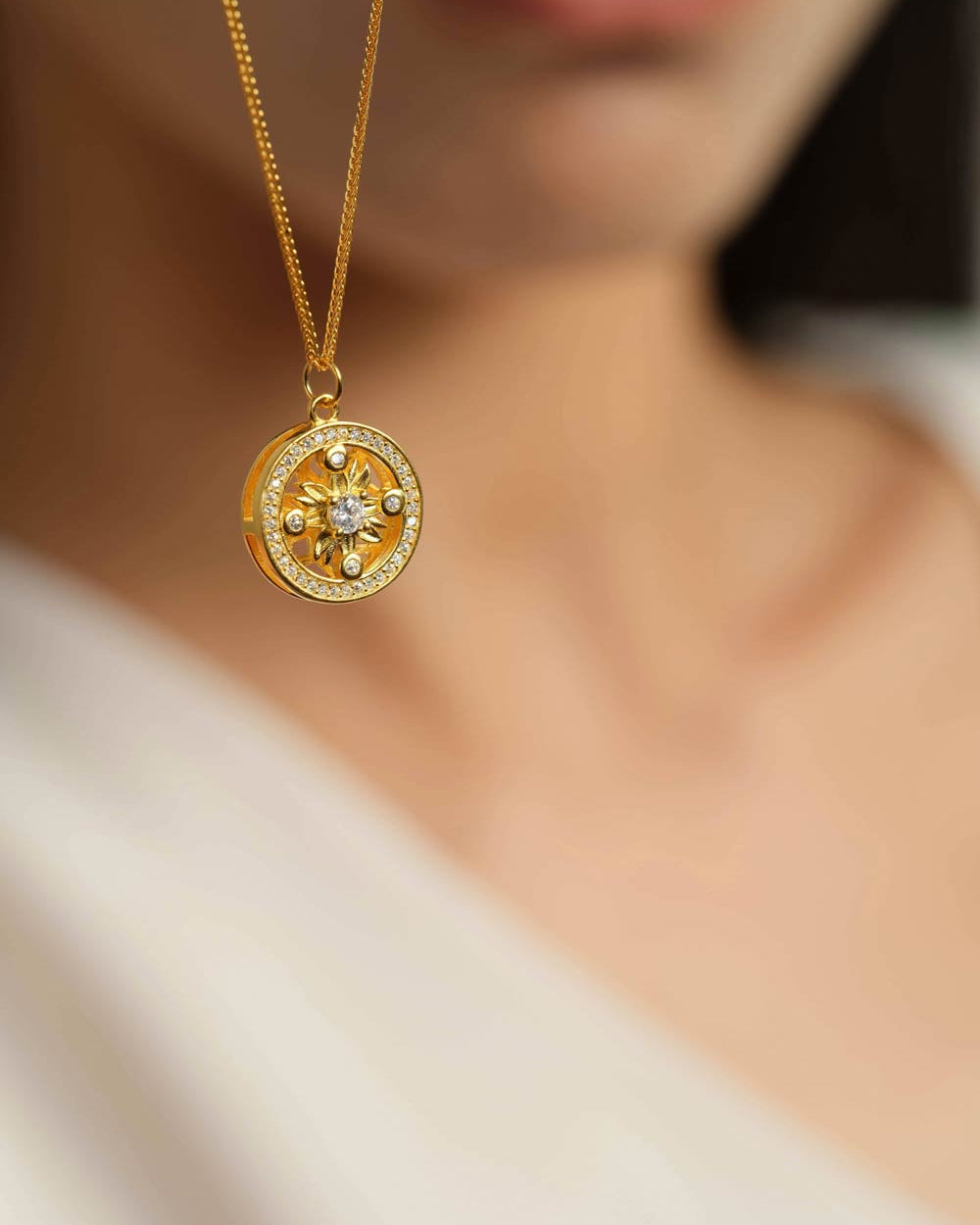 A gold pendant necklace with a circular design and small clear stones, worn by a model whose neck and lower face are softly blurred in the background.