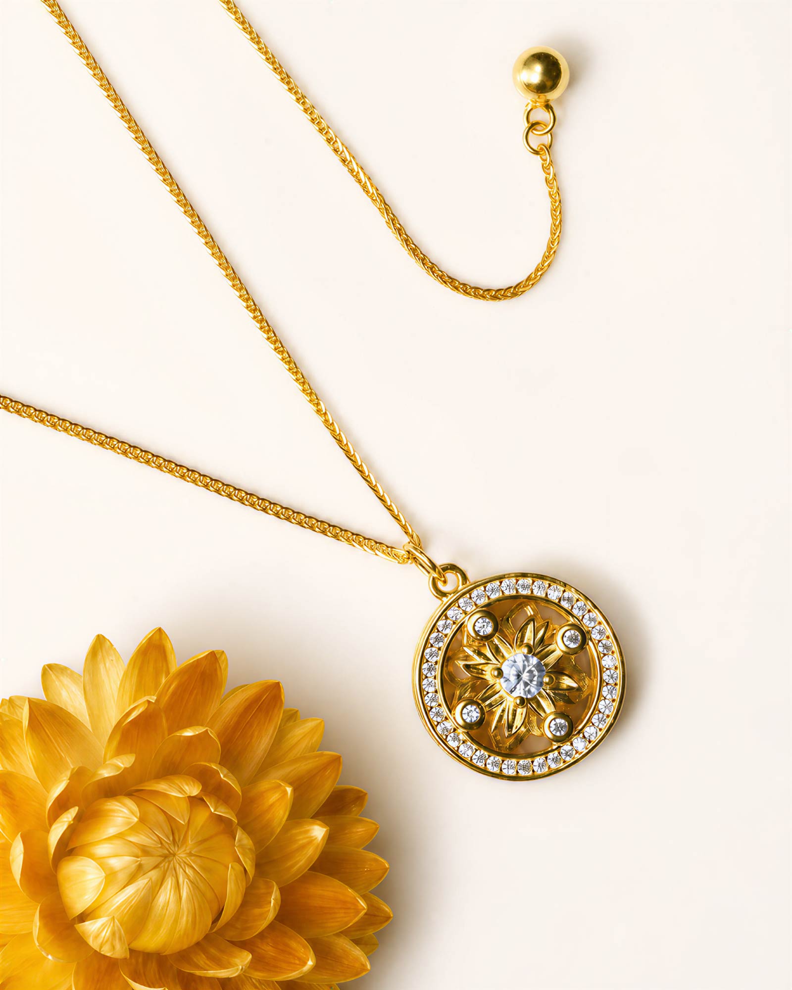A gold pendant necklace with a circular compass design, featuring a central clear gemstone surrounded by small clear stones, displayed on a light cream background next to a yellow flower.