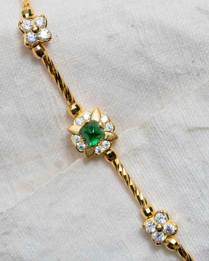A gold chain bracelet featuring three floral motifs, with a central green emerald-cut stone surrounded by small clear stones, set against a textured off-white fabric background.