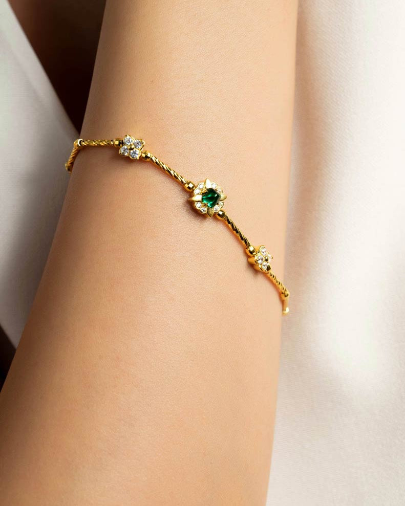 A delicate gold chain bracelet with a central green gemstone surrounded by small clear stones, worn on a person's wrist against a soft light background.