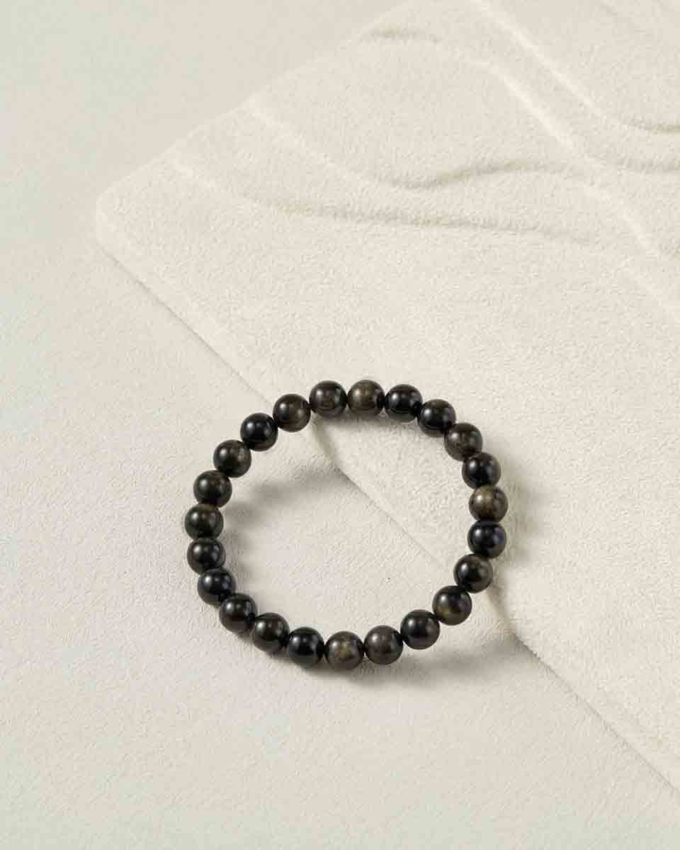 Still life photography of Choodo pure natural Gold Sheen Obsidian crystal beaded bracelet laid flat on an oatmeal velvet jewelry display board, capturing the minimalist premium textures.