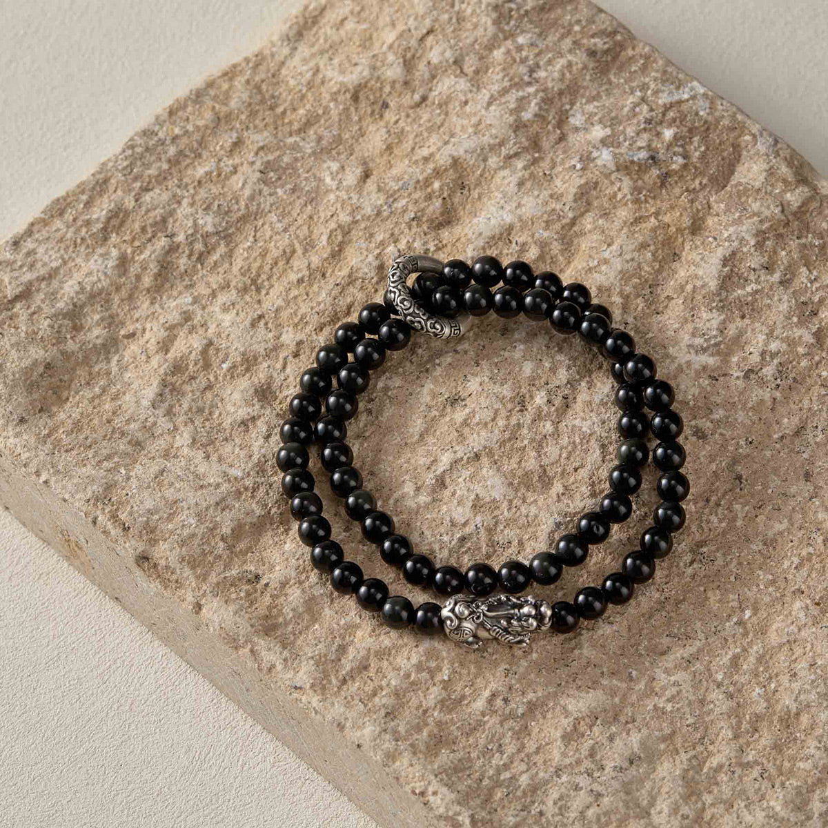 Still life photography of Choodo men's natural Obsidian bracelet with silver Pixiu laid flat on a rustic natural stone board, organic masculine textures contrast.
