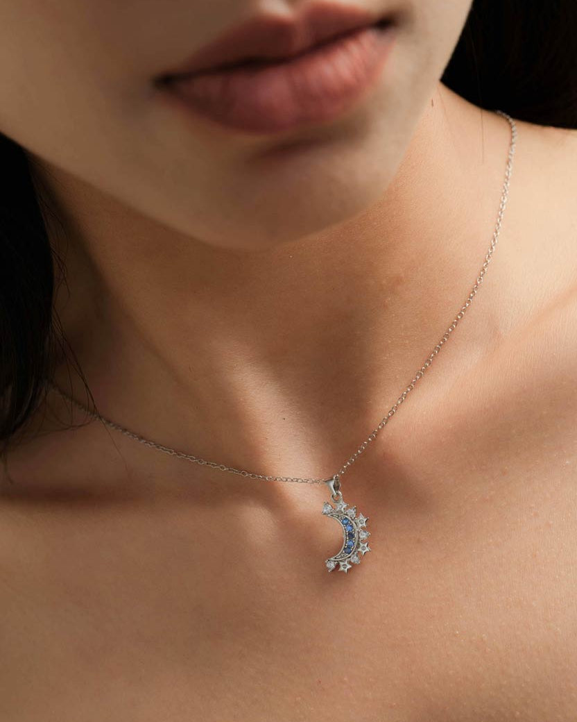 A close-up of a model's neck and chest wearing a delicate silver chain necklace with a crescent moon pendant adorned with small blue and clear stones.