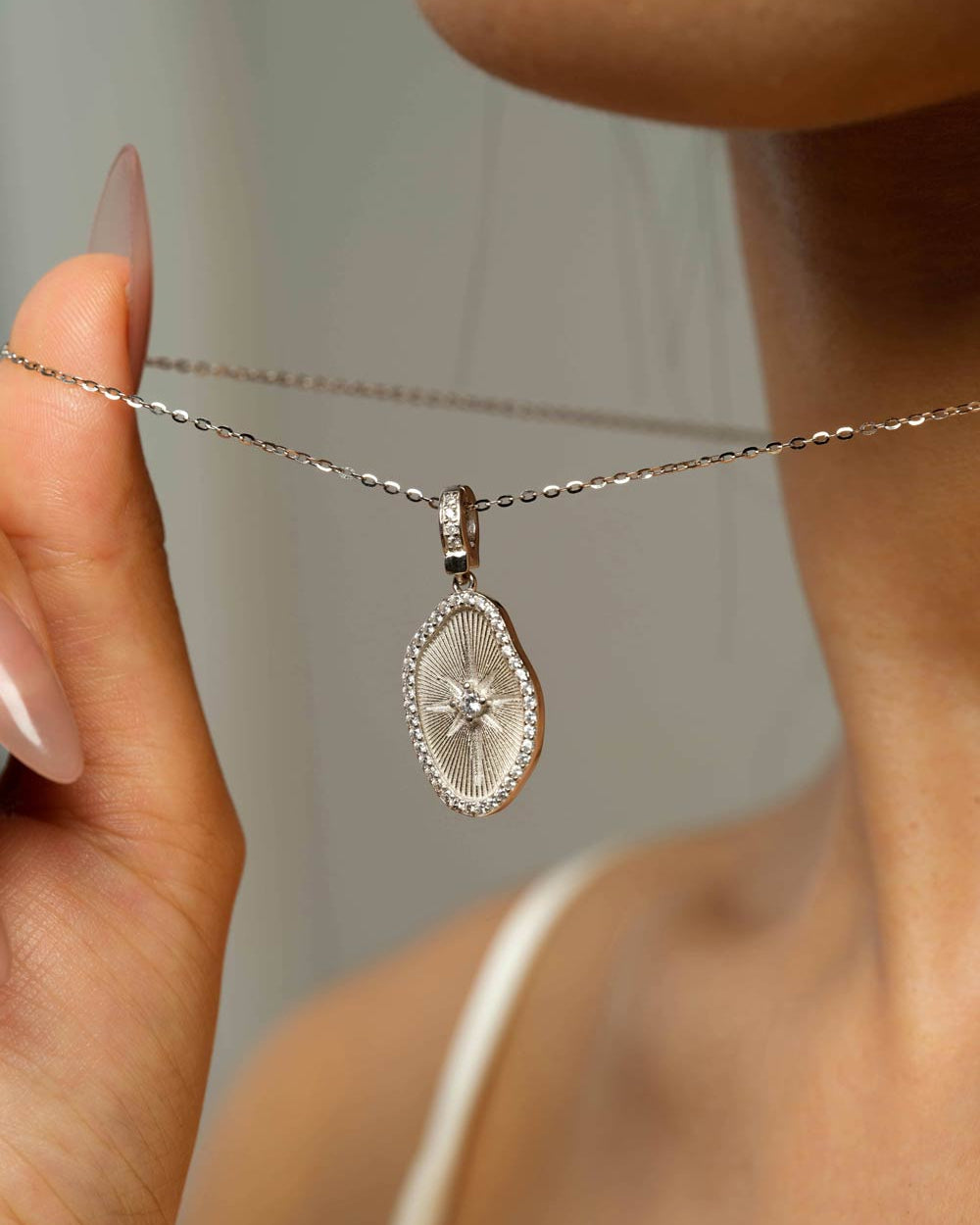A close-up shot of a model wearing a silver oval pendant necklace with a sunburst star design in the center, held by a hand on the left.