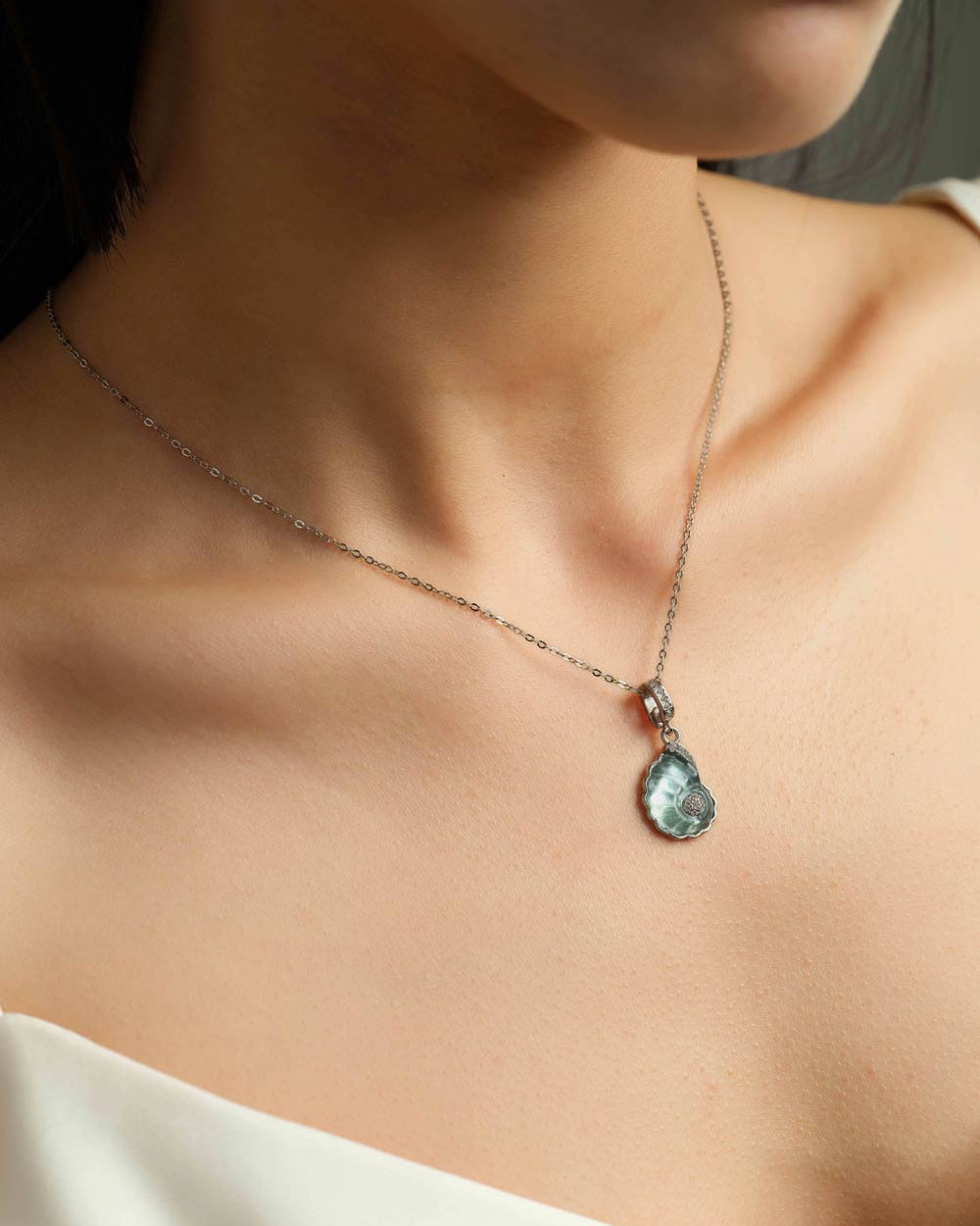 A close-up of a model's neck and chest wearing a delicate silver chain necklace with a shell-shaped light blue pendant.