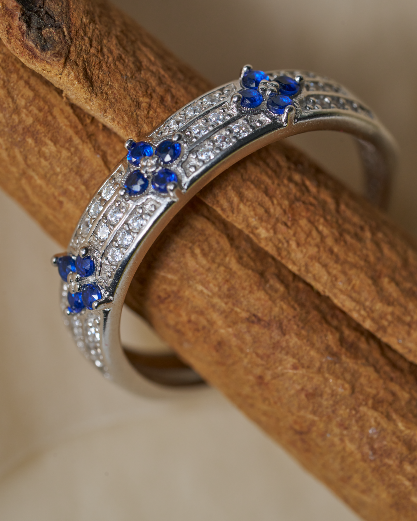 A silver-toned curved ring with rows of small clear stones and three blue gemstone floral motifs, displayed on a textured brown wooden stick against a soft beige background.