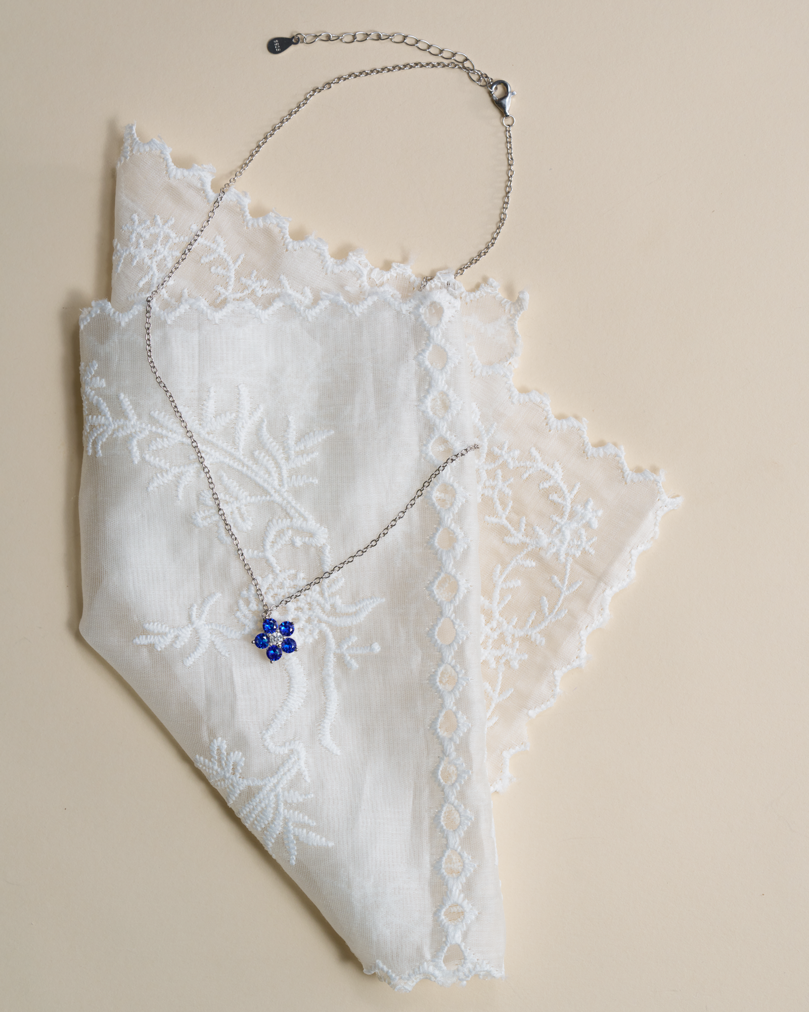 A silver chain necklace with a small blue flower-shaped pendant, displayed on a white embroidered lace fabric against a warm beige background.