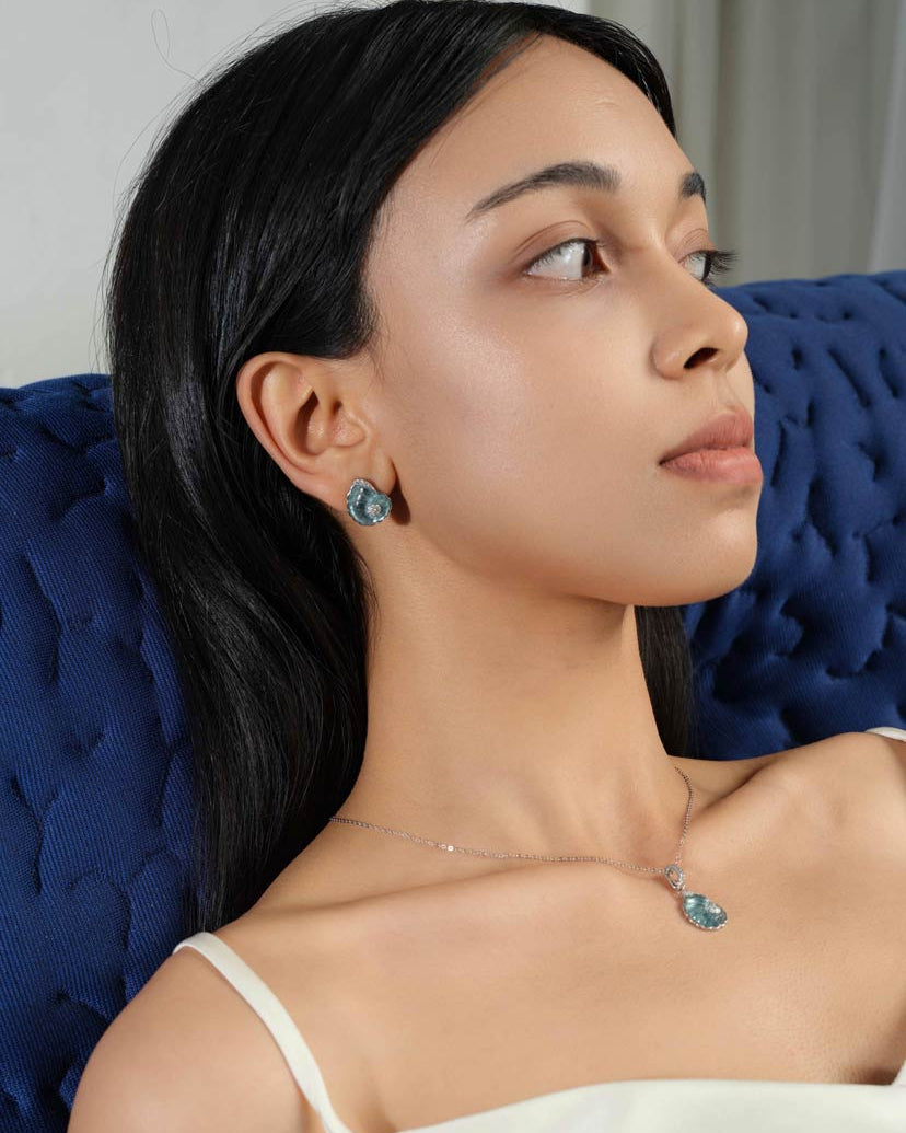 A woman wearing a delicate silver necklace and matching stud earring with a small blue shell-shaped pedants, posed against a dark blue tufted chair.