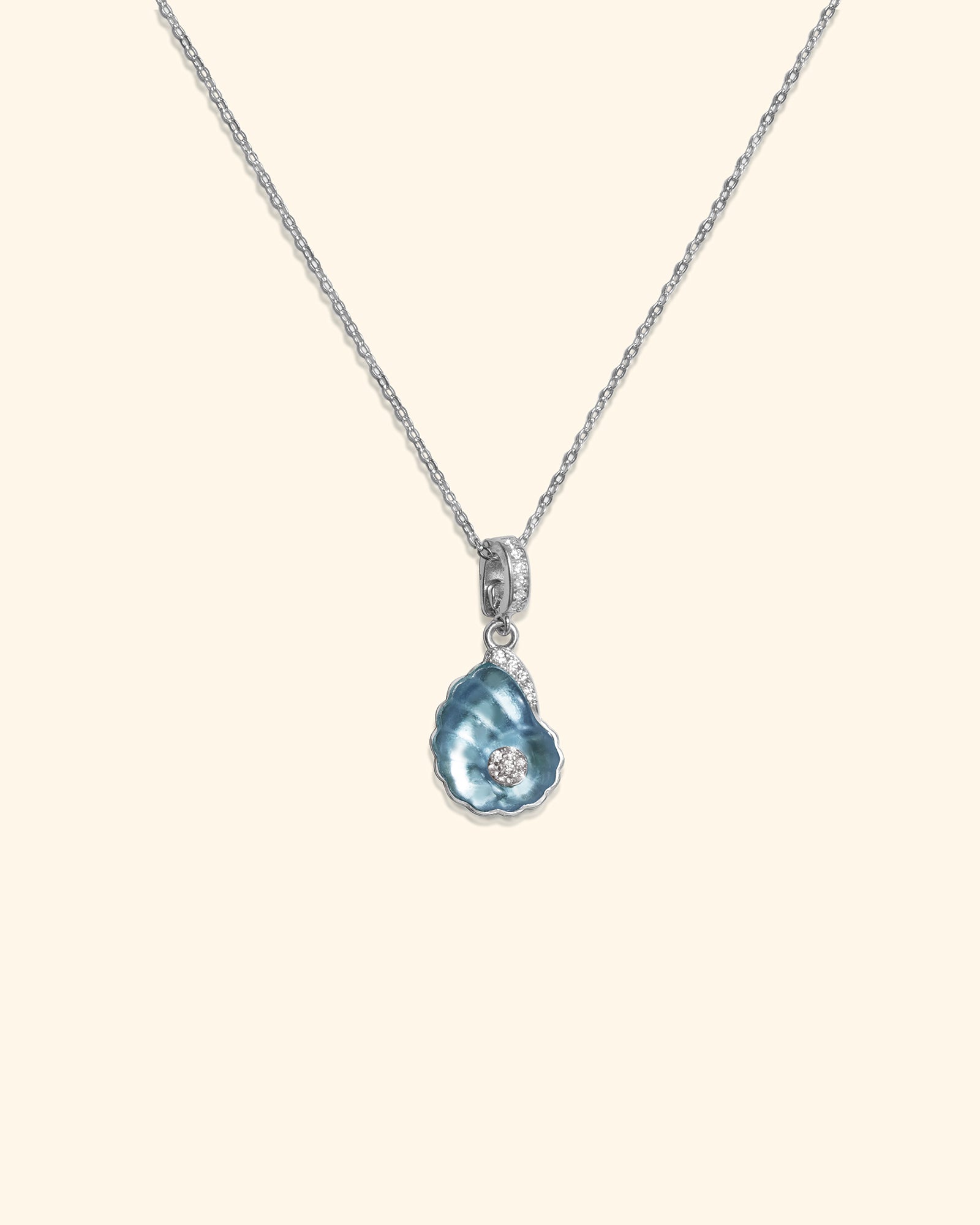 A delicate silver chain necklace featuring a blue shell-shaped crystal pendant with a light blue iridescent center and a small zircon, set against a solid cream background.